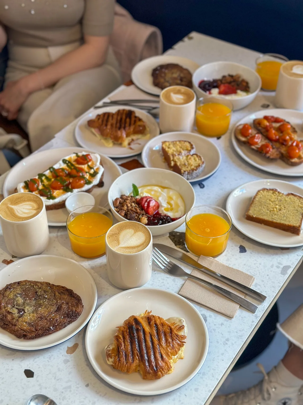 A table spread with coffees, orange juices, toasts, a granola and yoghurt bowl, a toasted croissant and cakes