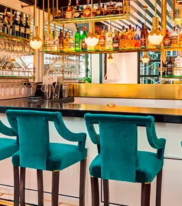A brass-topped bar counter with two teal velvet stools drawn up to it, backed by a lit spirits shelf and rows of hanging wine glasses