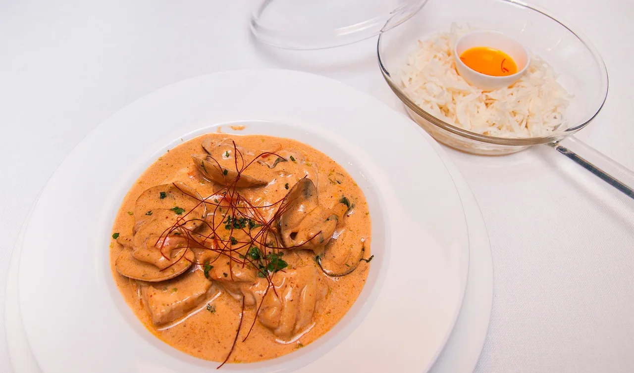 A white plate of stuffed pasta in an orange sauce with a small dish of egg yolk