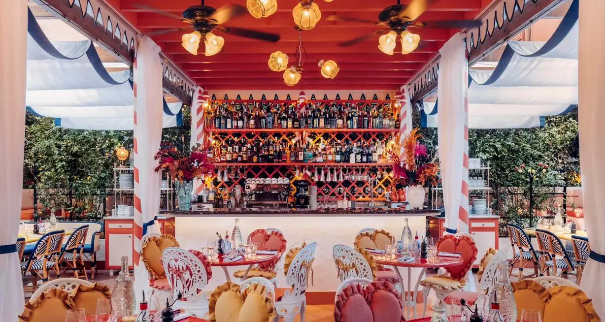 Vibrant covered terrace restaurant with a red ceiling, ornate glass lanterns, a well-stocked back bar with lattice shelving and small round tables ringed by white metal chairs with ruffled yellow and coral cushions