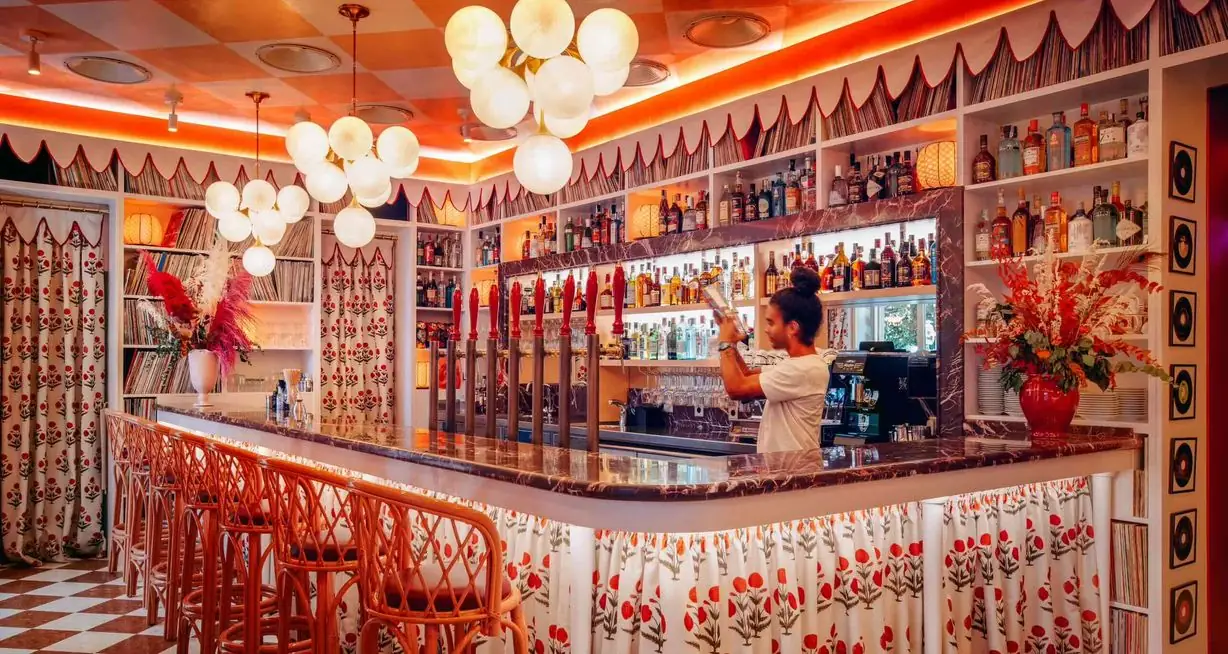 Retro cocktail bar with a marble-topped counter, red beer taps, walls lined with vinyl records and floral curtains, globe cluster chandeliers and a bartender mixing a drink behind the bar