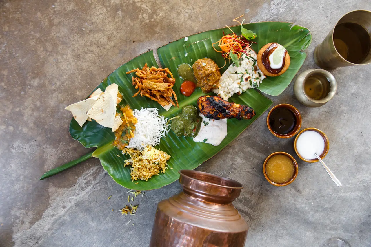 Indian thali on a banana leaf with tandoori chicken, chickpea curry, papadum, rice, biryani, and chutneys, served with a copper jug, at Bembi