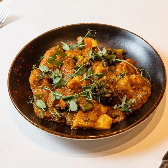 A dark speckled bowl on a cream tablecloth holding a mixed vegetable curry - cauliflower, broccoli, green beans, carrot and paneer in a deep orange sauce - scat