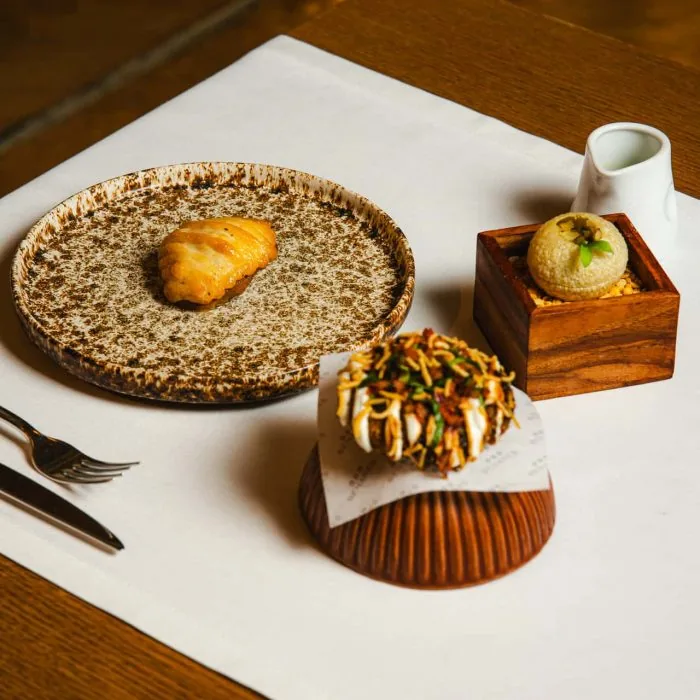 Three snacks on a white linen mat: a golden fried pastry alone on a speckled brown ceramic plate, a pale green sphere in a small wooden box beside a white jug,