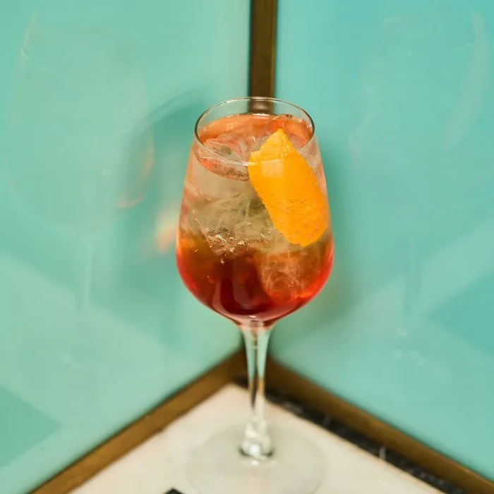 A stemmed wine glass of a red-orange spritz over ice with a curled orange peel, standing on a marble ledge against a turquoise tiled wall.