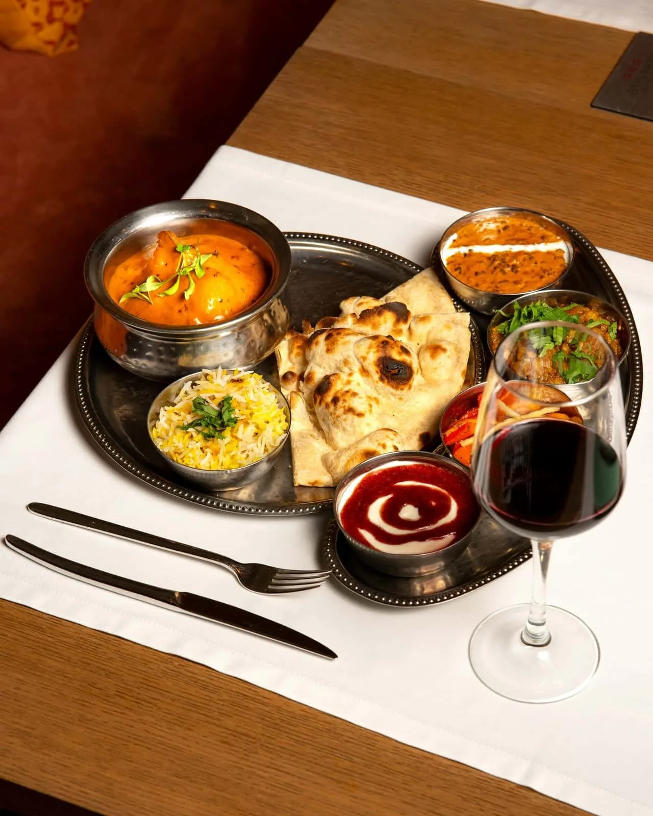 A metal thali tray with a bowl of orange curry, rice, naan bread and several small bowls of sauces beside a glass of red wine