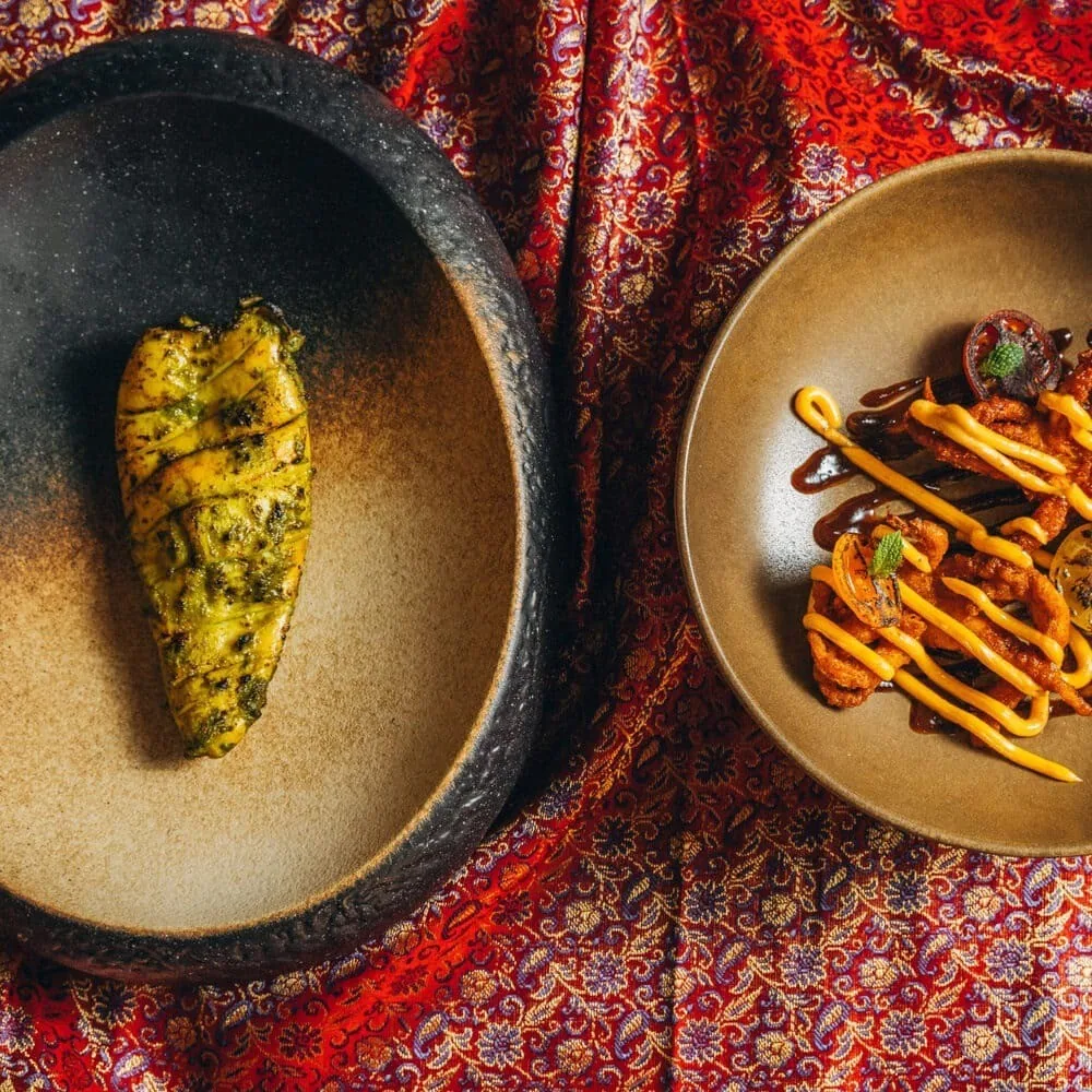 Two plates seen from above, one with a single green-marinated grilled piece and one with fried strips drizzled in yellow sauce, on red patterned fabric