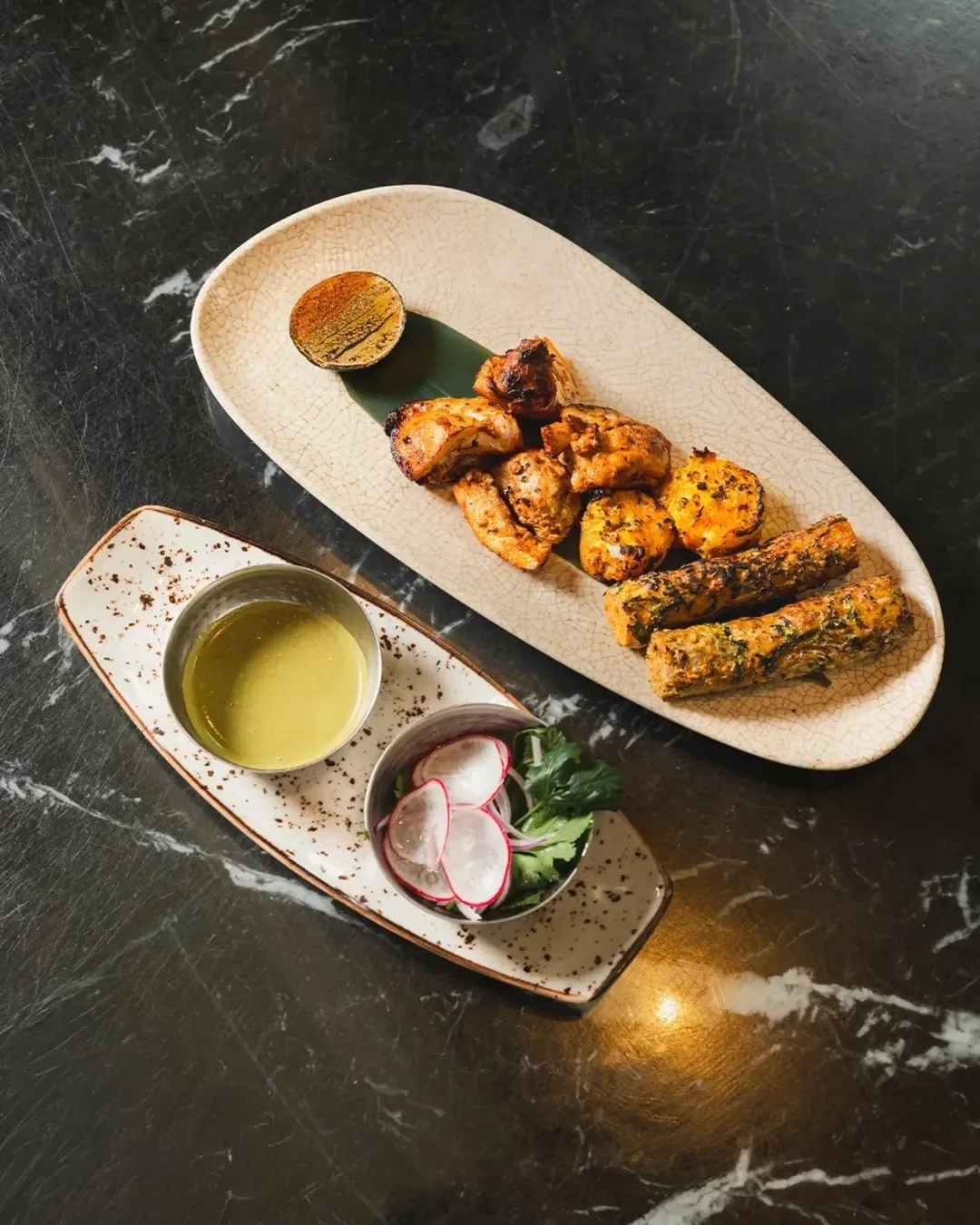 Grilled chicken pieces and a skewered kebab on a long serving plate beside small dipping sauces