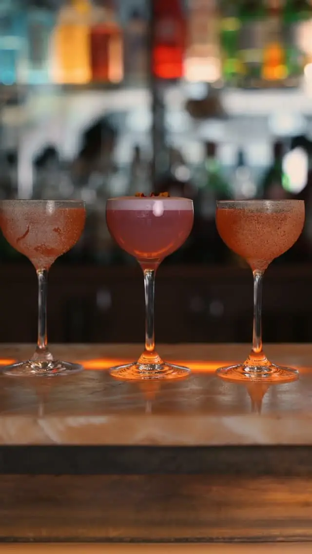 Three cocktails in coupe glasses lined up on a bar counter, backlit by bottle shelves.