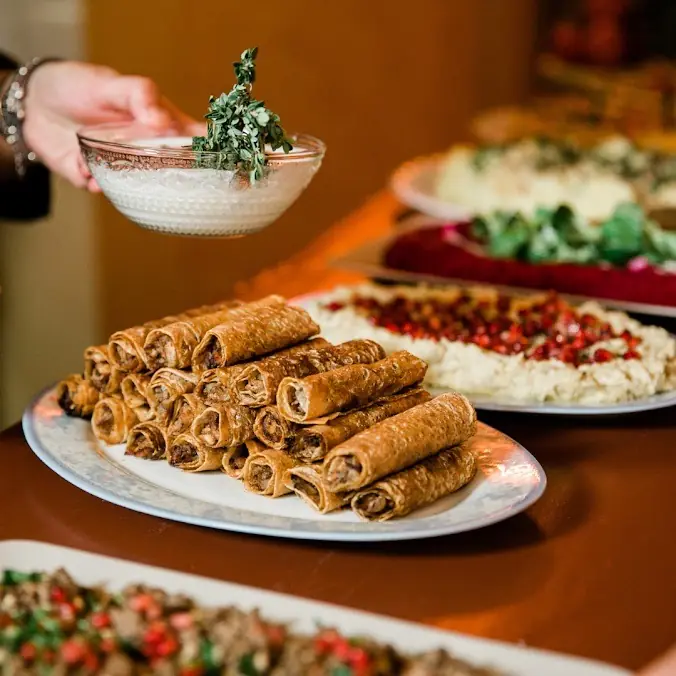 Stacked crispy rolled pastries beside plates of hummus topped with pomegranate seeds and greens, at Bērytī