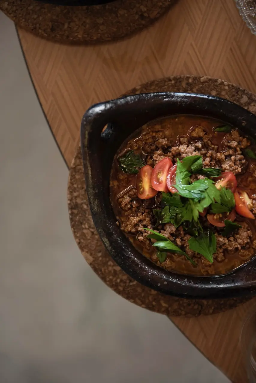 Overhead view of a dark clay dish of minced meat in broth topped with cherry tomatoes and parsley, at Bērytī