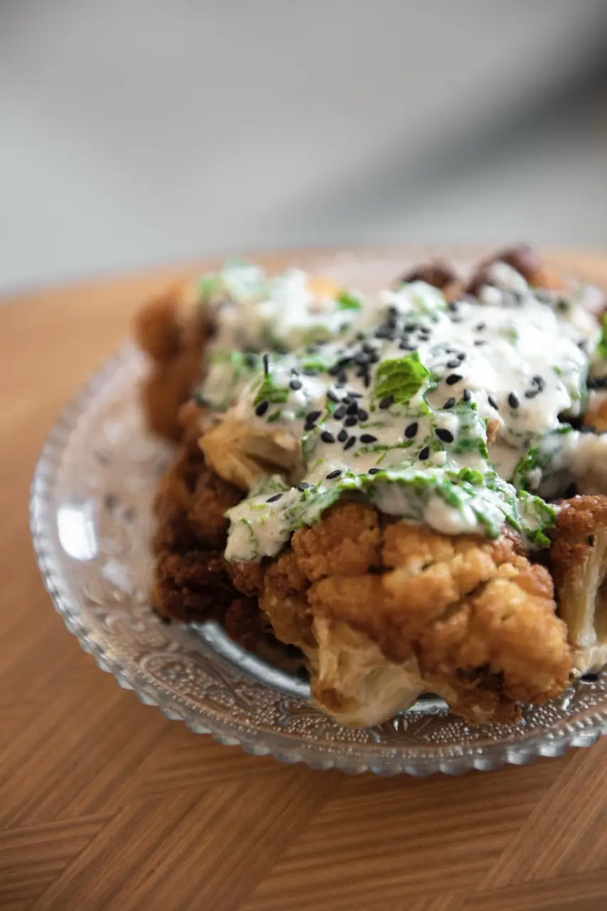 Fried cauliflower topped with a herbed yogurt sauce, black sesame seeds, and chopped greens on a glass plate, at Bērytī