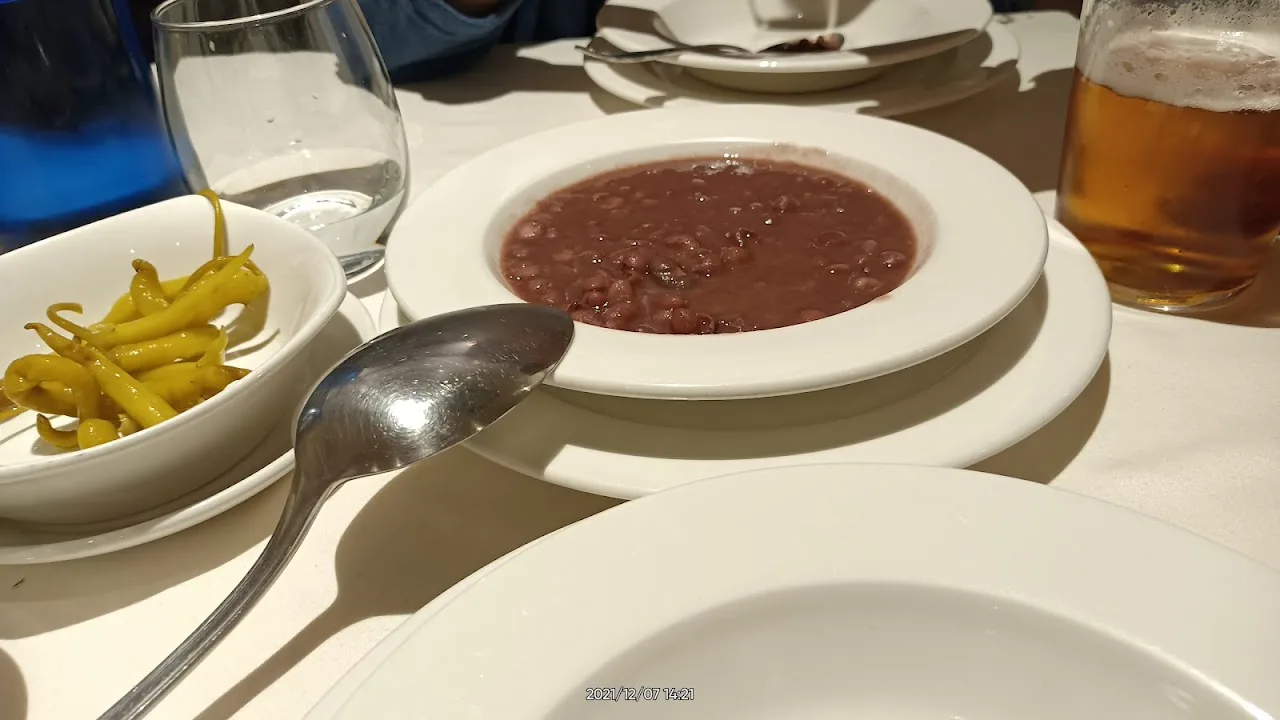 A bowl of dark bean stew served with a side of yellow beans and a beer