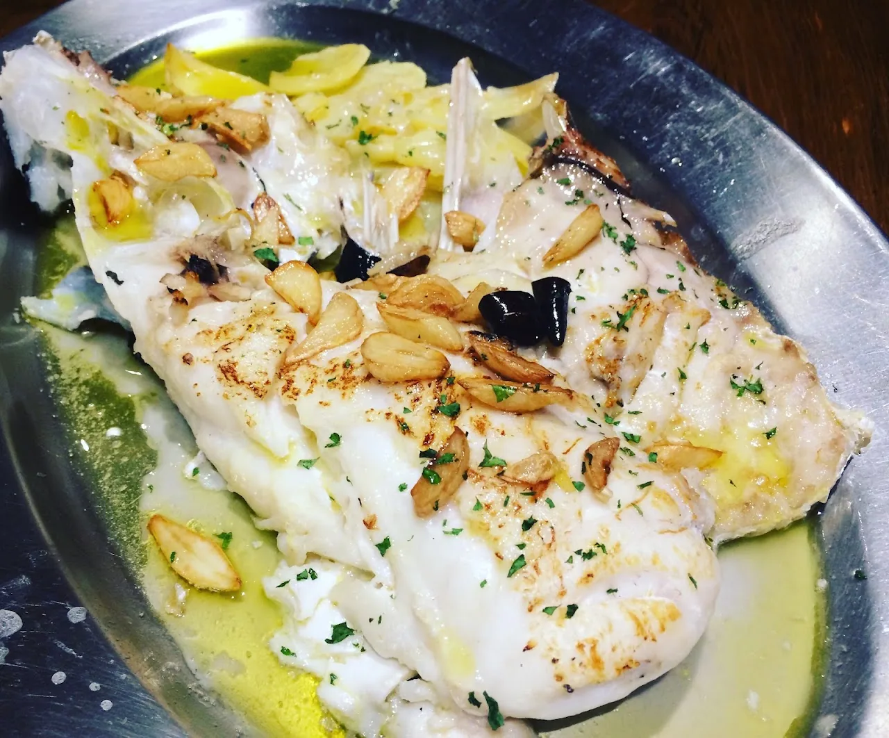 A whole grilled fish topped with sliced garlic on a metal platter