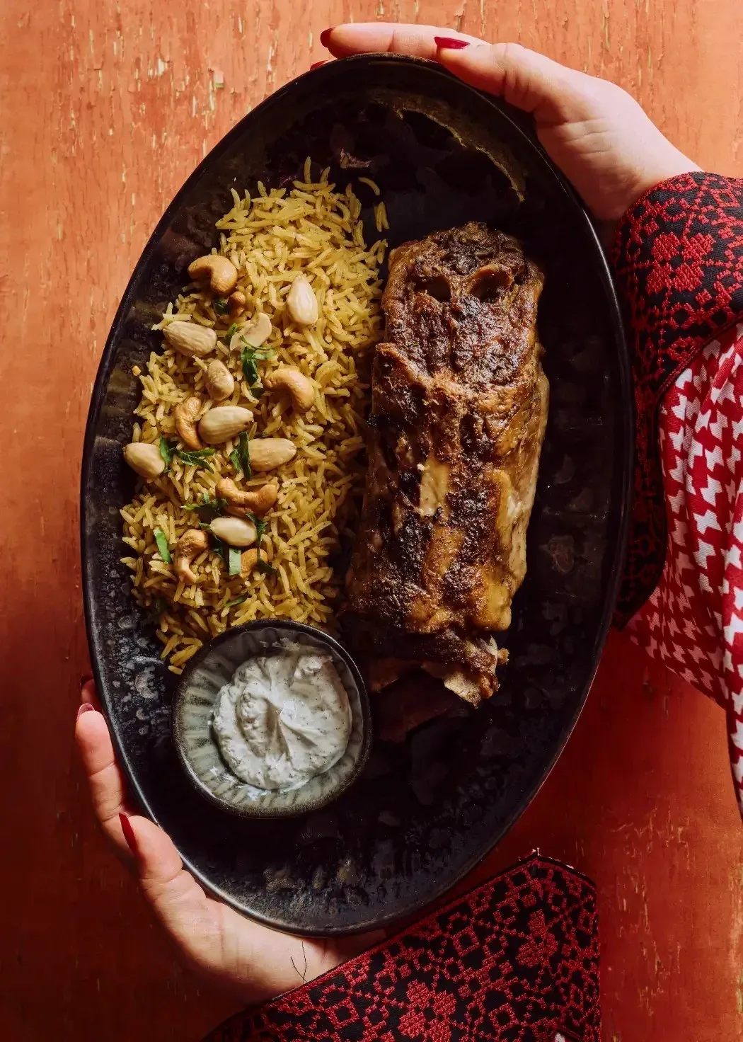 An oval dish of rice topped with meat and nuts held out in two hands