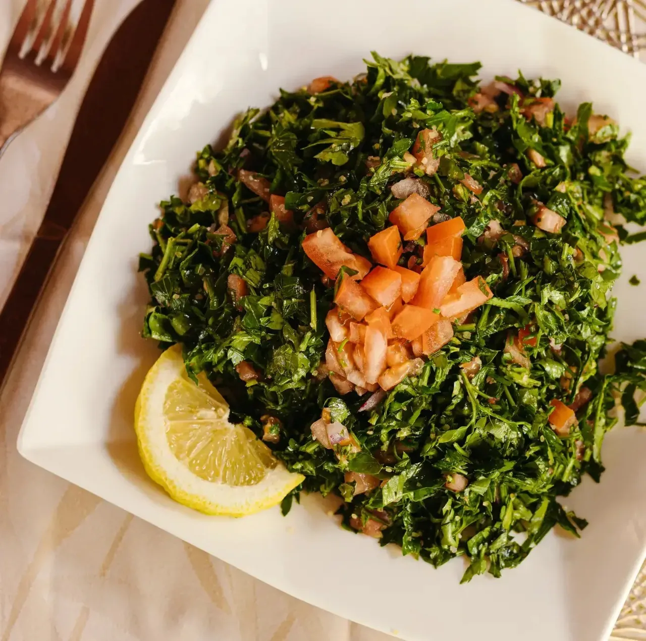 A square white plate of chopped parsley salad with tomato and a lemon wedge