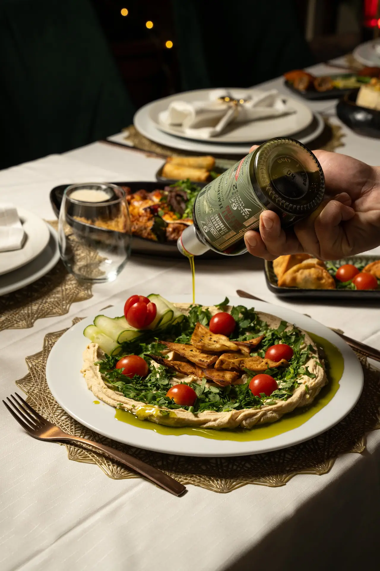 Hand drizzling olive oil over a hummus plate topped with parsley, cherry tomatoes, cucumber and pita chips