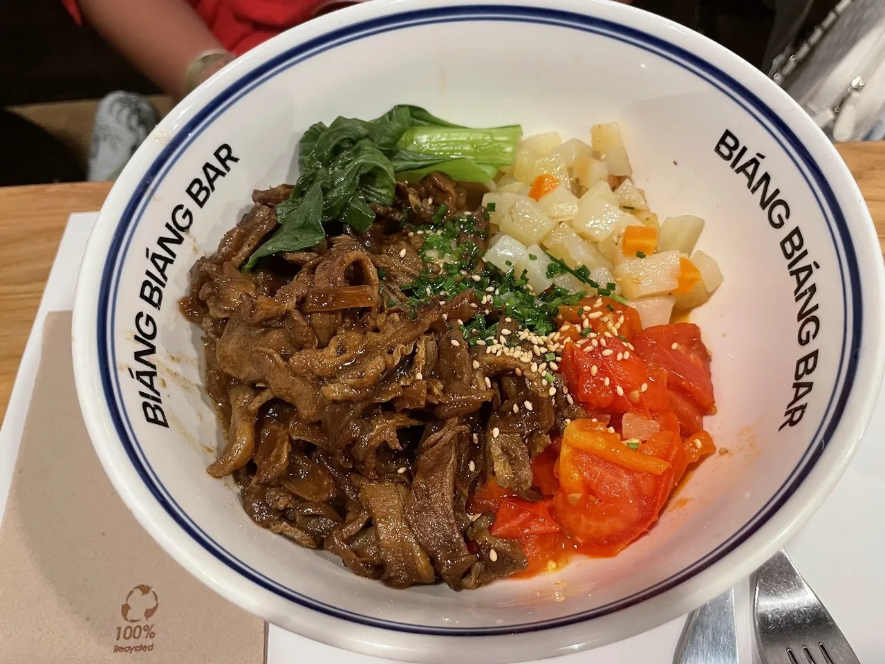 A white bowl of braised beef noodles with greens, tomato and diced vegetables