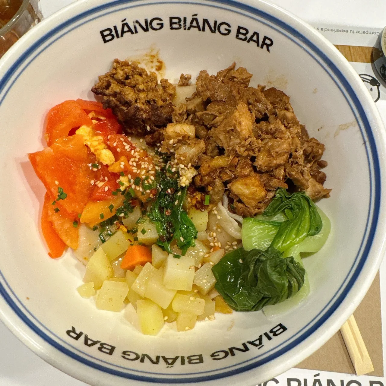A bowl of noodles topped with tomato, meat and greens