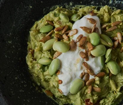 A dark stone bowl of guacamole topped with a spoonful of white cream, blanched broad beans and toasted sunflower seeds, shot from above