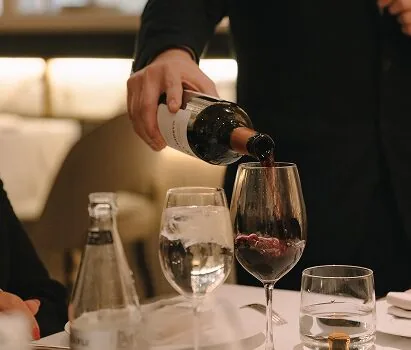 A waiter pouring red wine into a large stemmed glass at a white-clothed table, a bottle of water and a second glass alongside