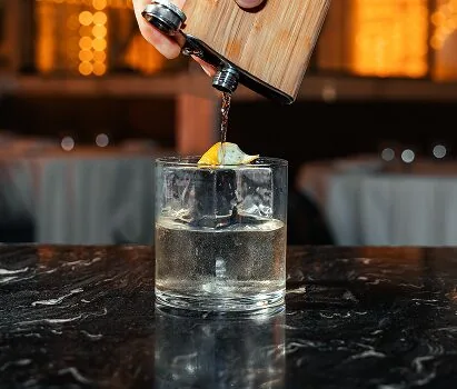 A cocktail being poured from a small wooden cask into a rocks glass over clear ice with a citrus twist, on a dark marble bar