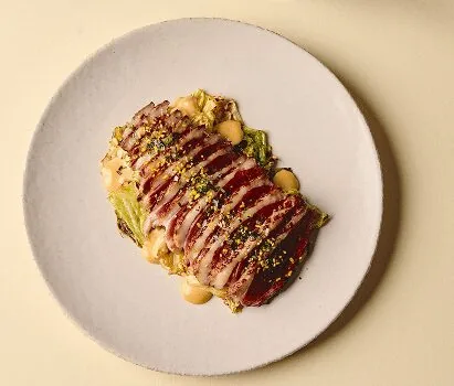 Slices of seared red tuna fanned over dressed leaves with dots of pale sauce and a scatter of chopped pistachio, on a round pale plate shot from directly above
