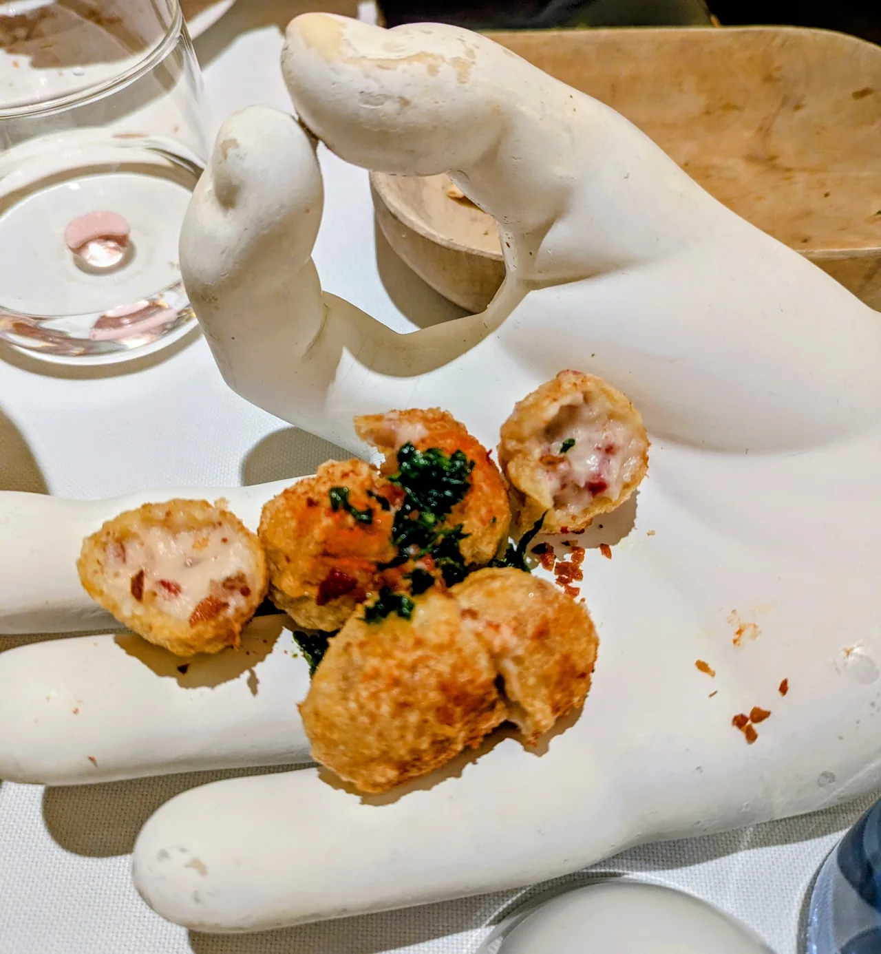 Fried croquetas, some cut open to show a creamy filling with pink flecks, on a white sculpted hand with green herb garnish