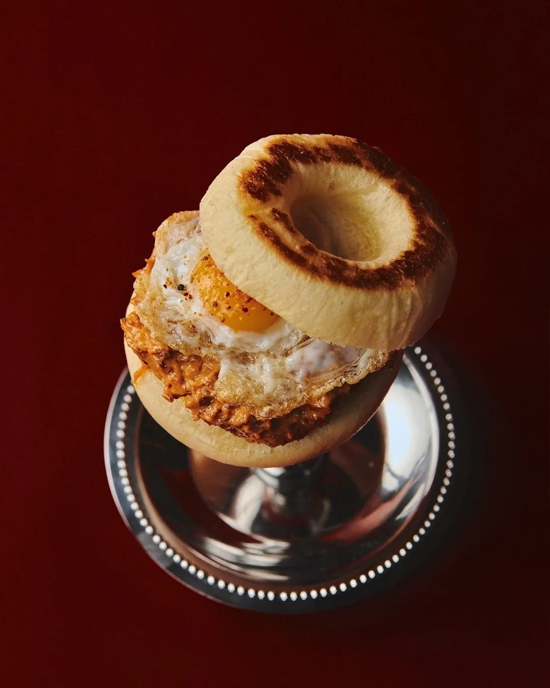A toasted brioche bun topped with a fried egg on a silver stand against a deep red background