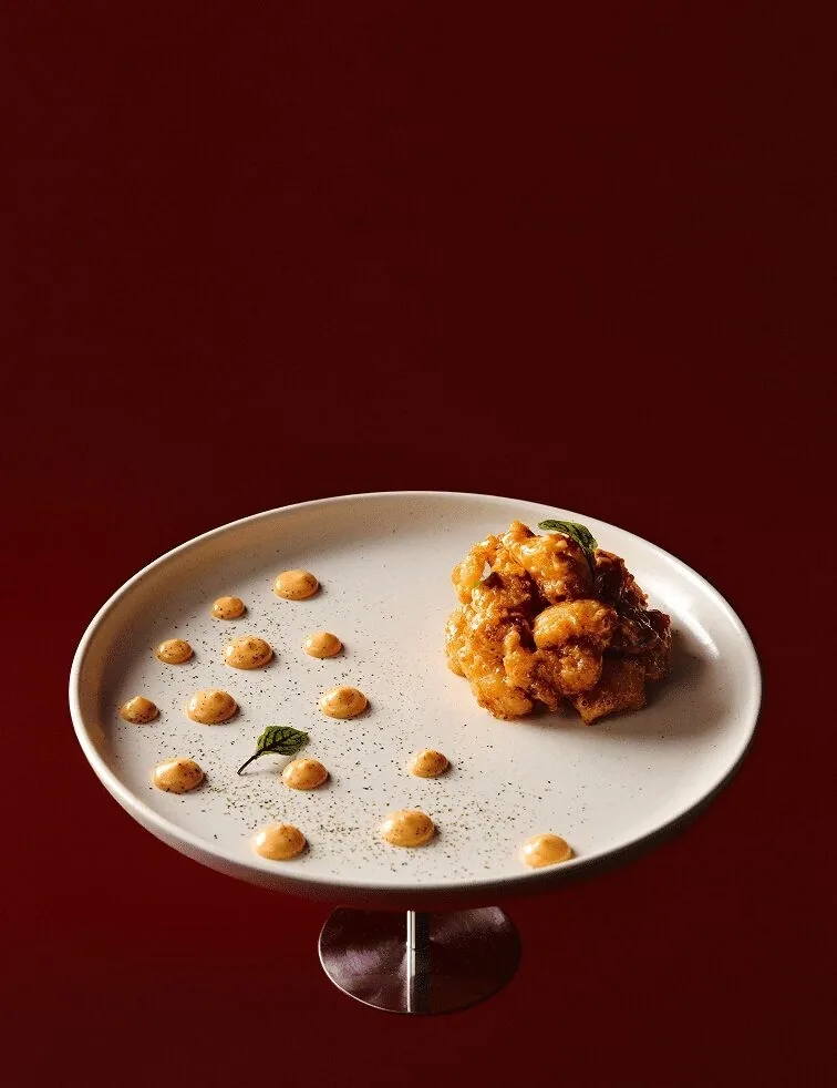 A white plate with a cluster of fried morsels and dots of orange sauce against a deep red background