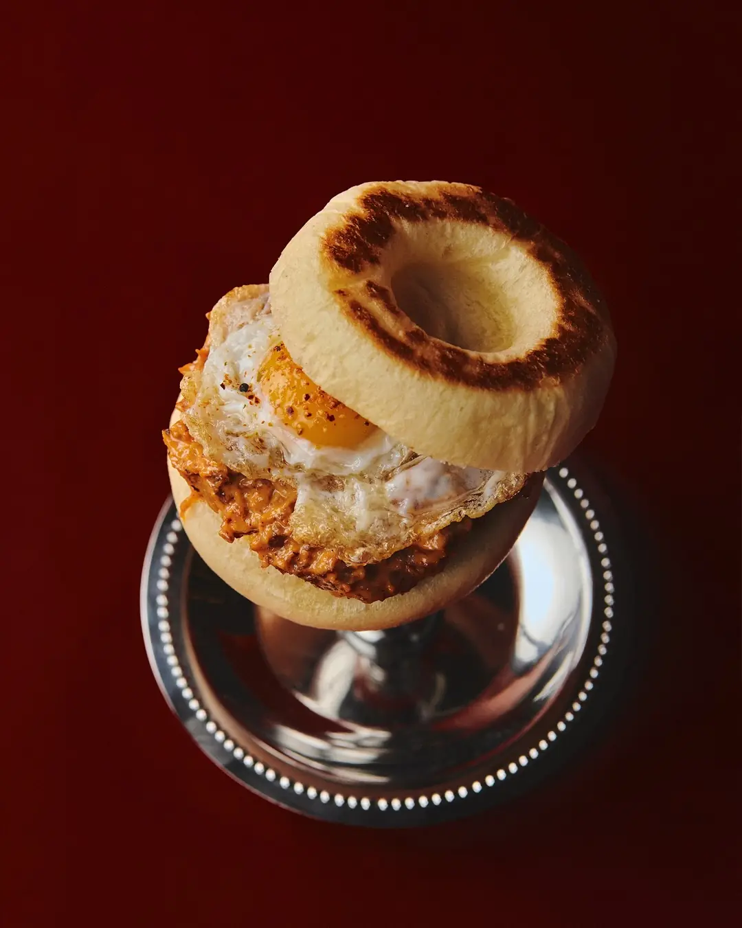 An English muffin sandwich with a fried egg and a spiced meat filling, served on a silver stand