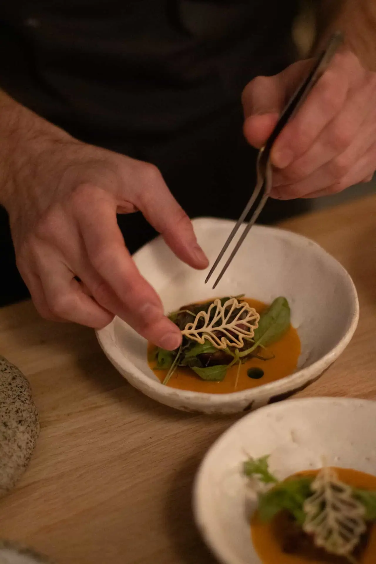 A cook uses long tweezers to set a lacy leaf-shaped cracker onto a bowl of orange sauce with green leaves and dark braised meat, a second identical bowl waiting on the wooden pass