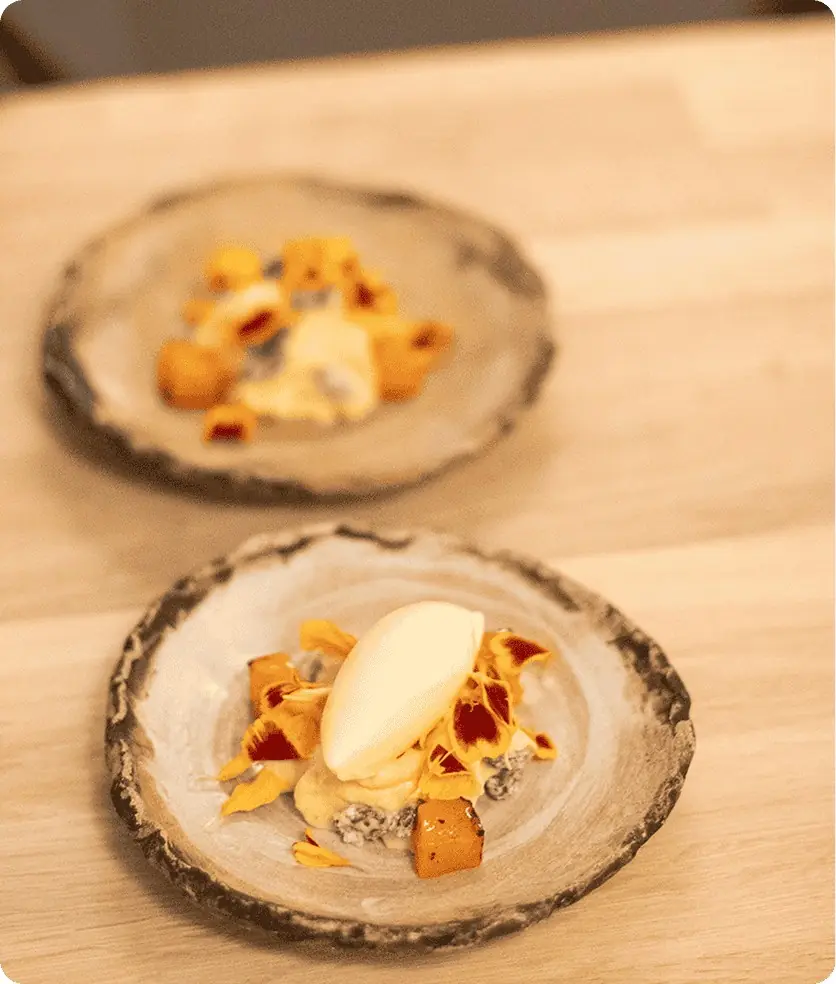 Two rough-edged ceramic plates on pale wood, each holding a quenelle of pale ice cream over orange marigold petals, cubes of glazed squash and a dark crumb