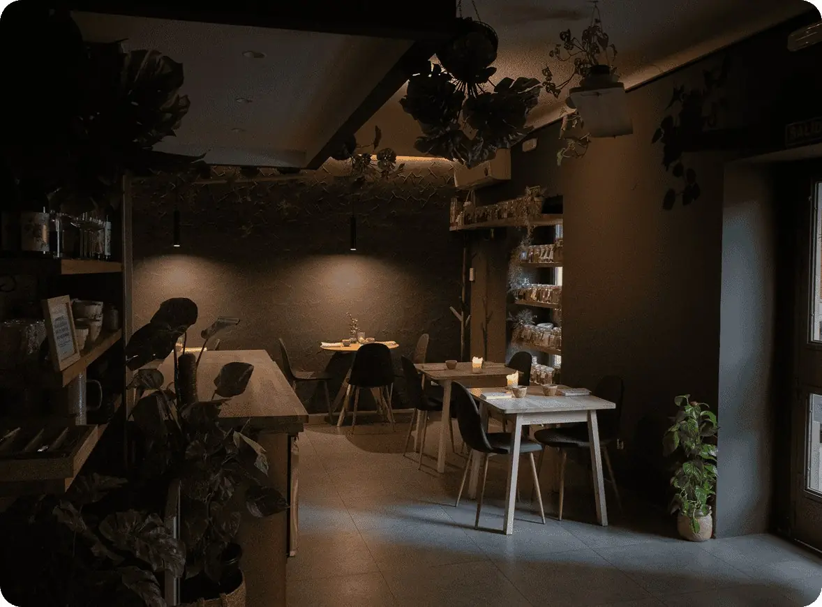 Small dim dining room with hanging and potted plants, a textured dark wall, open shelves of glassware and ceramics, and a few pale wood tables laid with small dishes and a candle