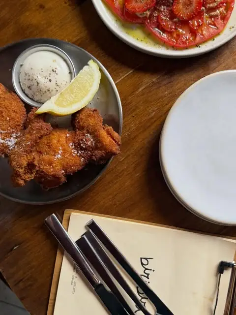 Breaded fried cutlets with a lemon wedge and pepper-flecked aioli on a metal plate, a sliced tomato salad behind, on a dark wooden table