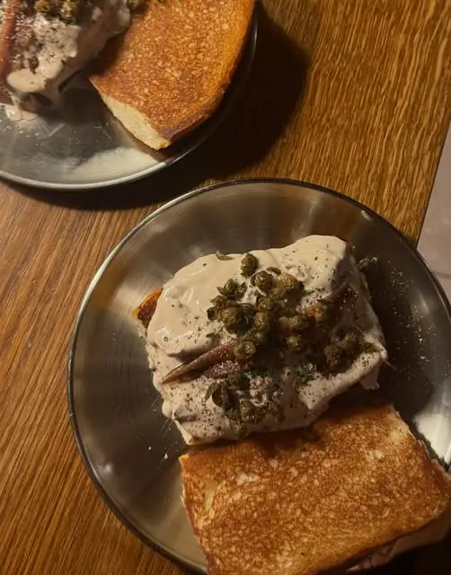 Toasted bread topped with anchovy fillets, fried capers and a cream sauce on metal plates set on a wooden table