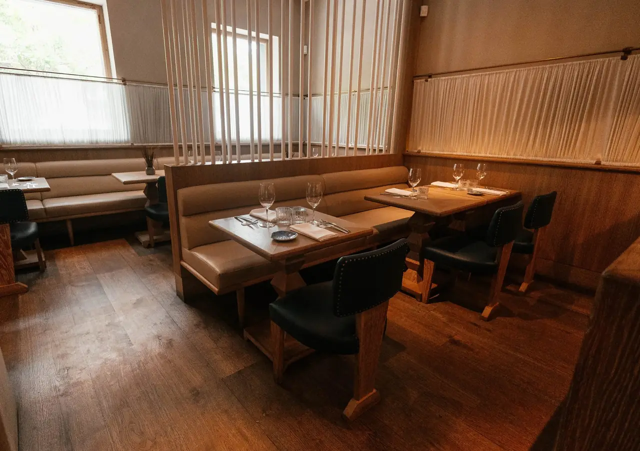 Dining room with cream leather banquettes, oak tables laid with wine glasses and folded napkins, slatted wood screens and a pleated fabric wainscot along the wall