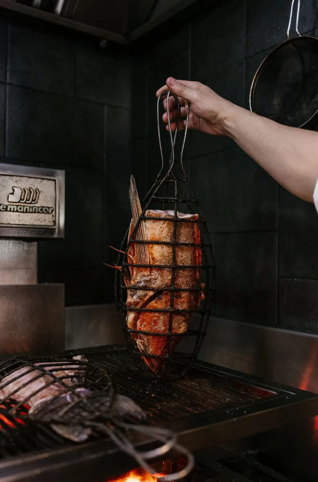 A cook's hand lowering a whole fish clamped in a wire grill basket over glowing charcoal, a second fish already searing on the grate below