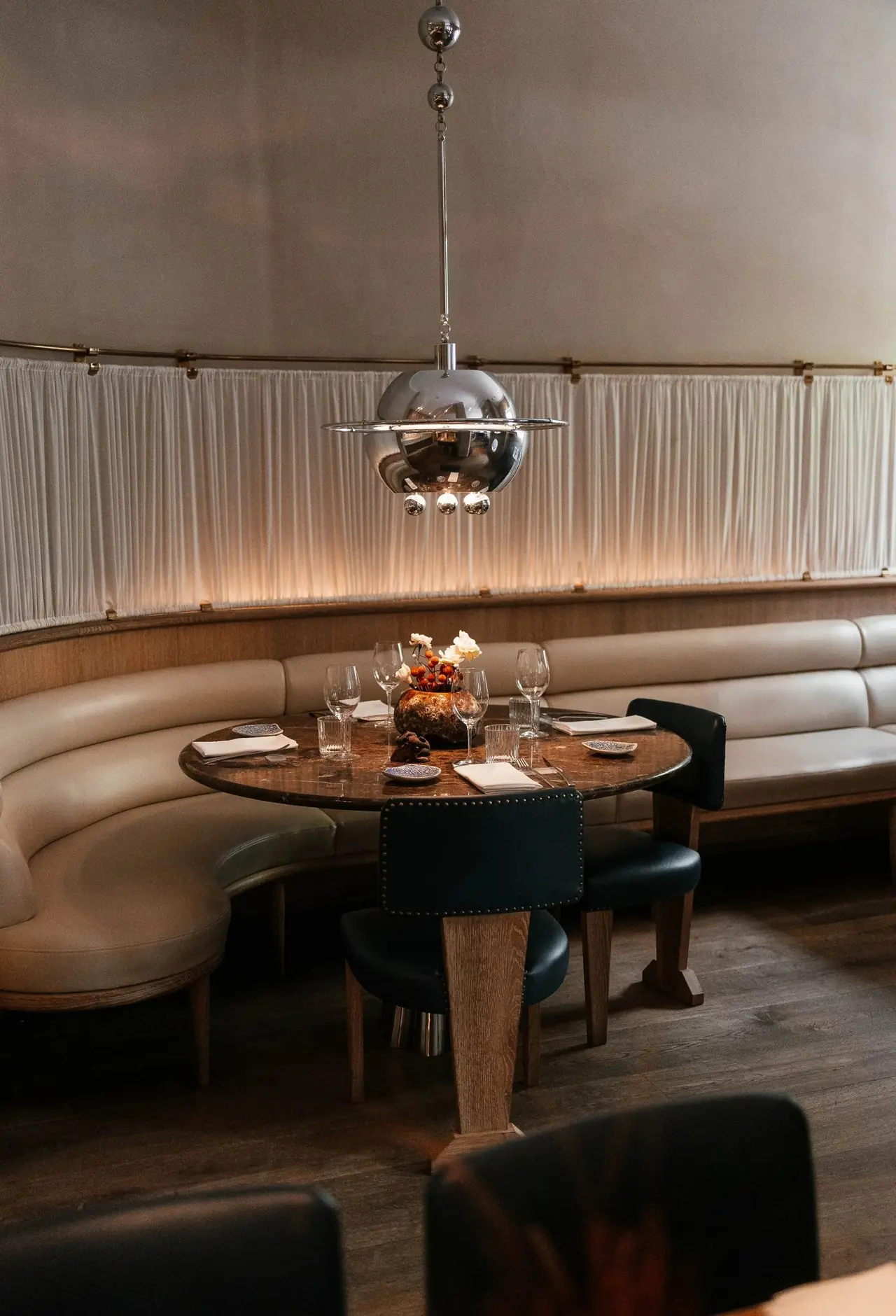 Round marble table set for four in a curved cream banquette booth, chrome globe pendant lamp overhead and a backlit pleated curtain wall behind