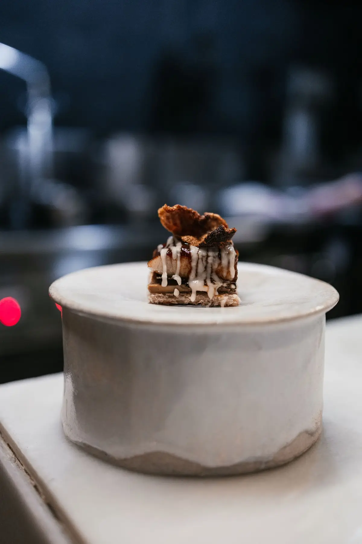 Small plated bite with sauce drizzle and a crisp wafer on top, served on a tall ceramic pedestal