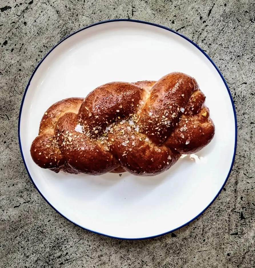 A braided challah bread topped with seeds and flaky salt, served on a white plate with a blue rim, Bistrot Levante