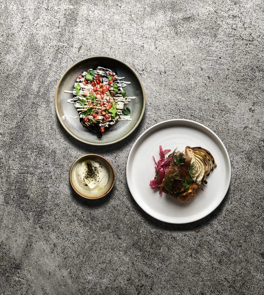 Roasted eggplant with pomegranate seeds and yogurt drizzle next to a small dish of labneh and a plate of grilled meat with pickled onions and herbs, Bistrot Levante