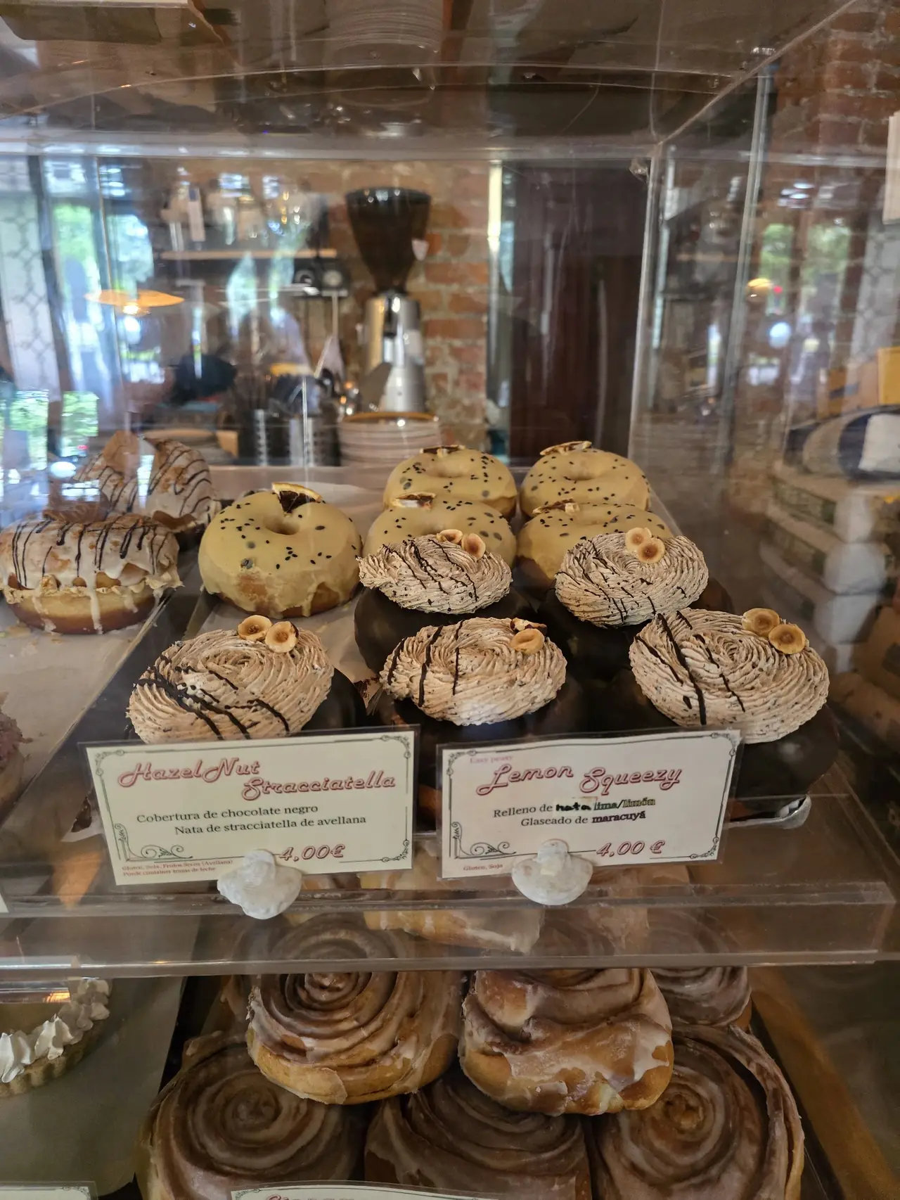 Bakery display case of glazed donuts and cream-topped pastries labeled Hazelnut Stracciatella and Lemon Squeezy
