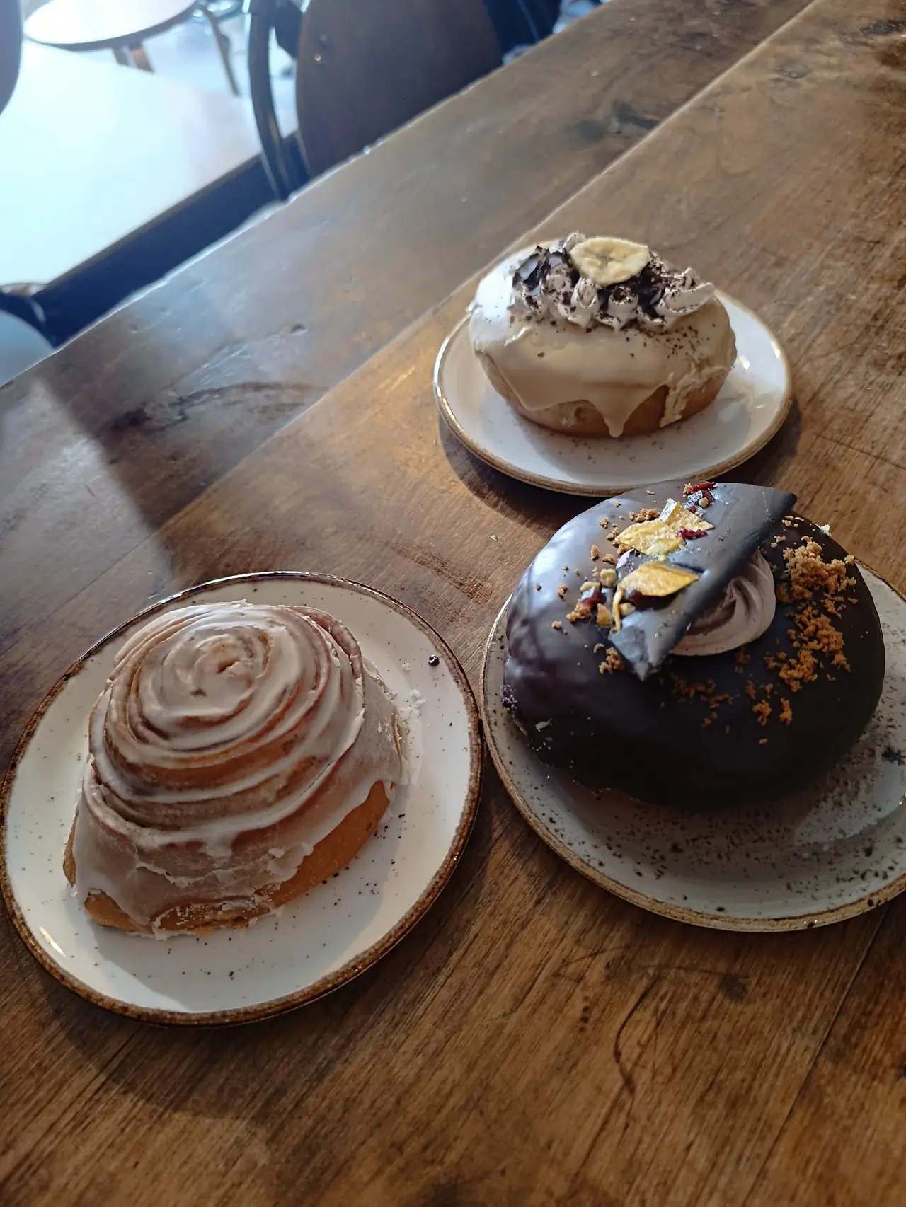 Three plated pastries on a table: a cinnamon roll, a banana cream donut and a chocolate-glazed donut with gold leaf
