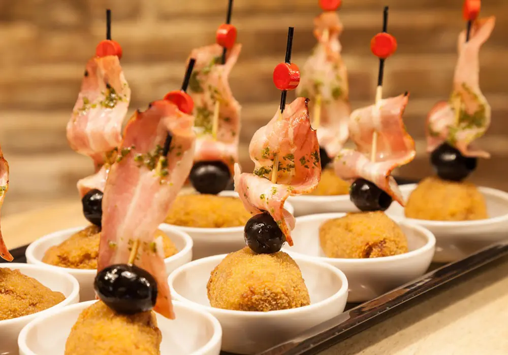 Fried croquettes topped with a black olive and a strip of cured ham on a skewer, served in small white bowls, at Blai 9