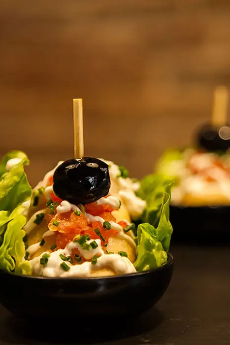 A pintxo of diced tomato, chives, and a black olive skewered atop bread with lettuce leaves, in a small dark bowl, at Blai 9