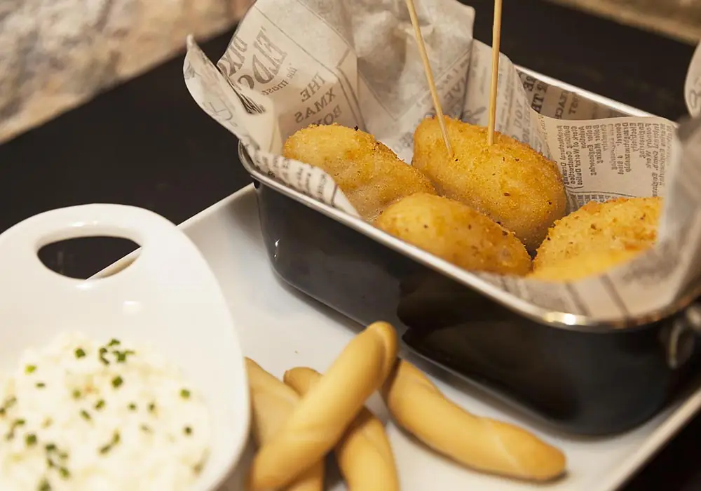 Breaded croquettes on skewers in a newspaper-lined metal basket, with bread sticks and a bowl of aioli, at Blai 9