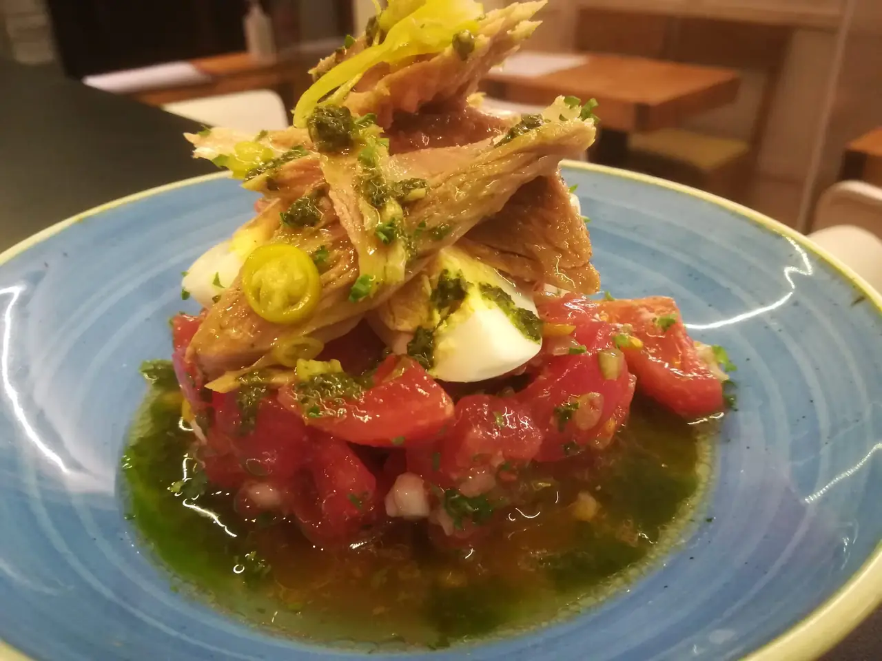 Layered starter of diced tomato, boiled egg, and crispy fish skin strips drizzled with green herb oil, on a blue ceramic plate at Blavis