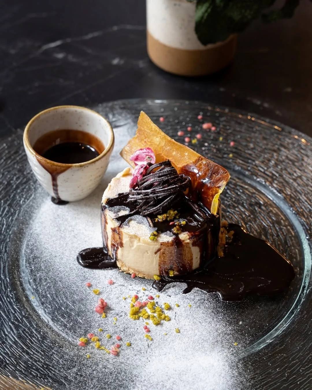 A dessert with chocolate strands and a small cup of coffee on a dark glass plate