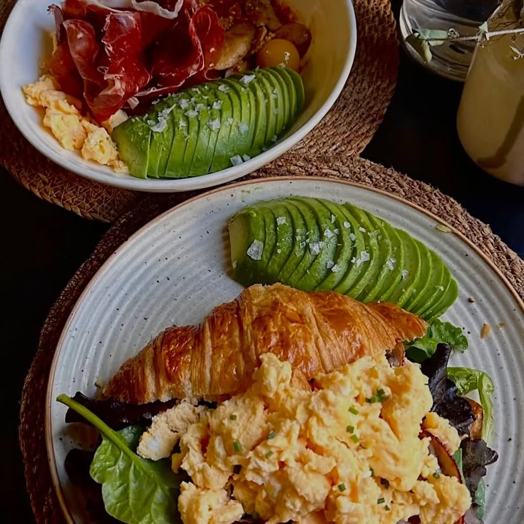 A plate with scrambled eggs, a croissant, sliced avocado and salad beside another bowl of eggs and tomato
